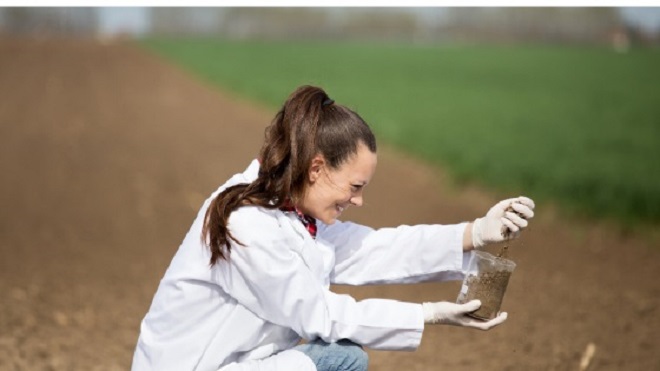 A young lady wearing white lab coat collecting a soil sample to a cleat glass jar.