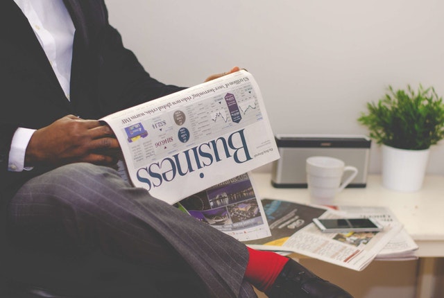 man-reading-newspaper-while-sitting-near-table-with-smartphone-and-cup