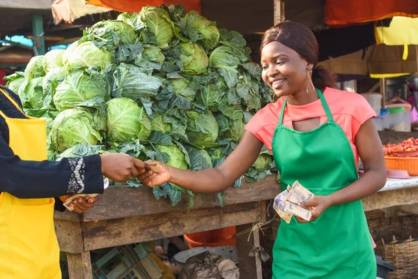 Happy African female farmer, trader, entrepreneur or businessman holding multiple cash notes in her hands serving a customer buying cabbages